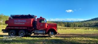 A red fire truck in a grassy field with trees and mountains in the background.