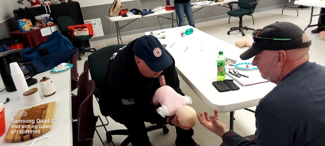 A firefighter is demonstrating CPR on a dummy to a man in a casual setting with tables and items around.