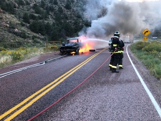 A firefighter approaching a burning car on a road.