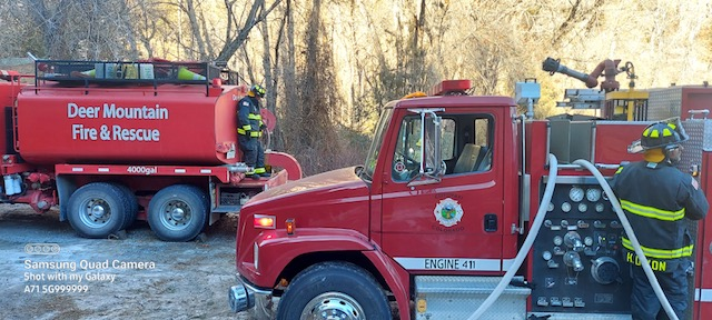 Firefighter by a fire engine, with a water tanker truck from Deer Mountain Fire & Rescue in the background.