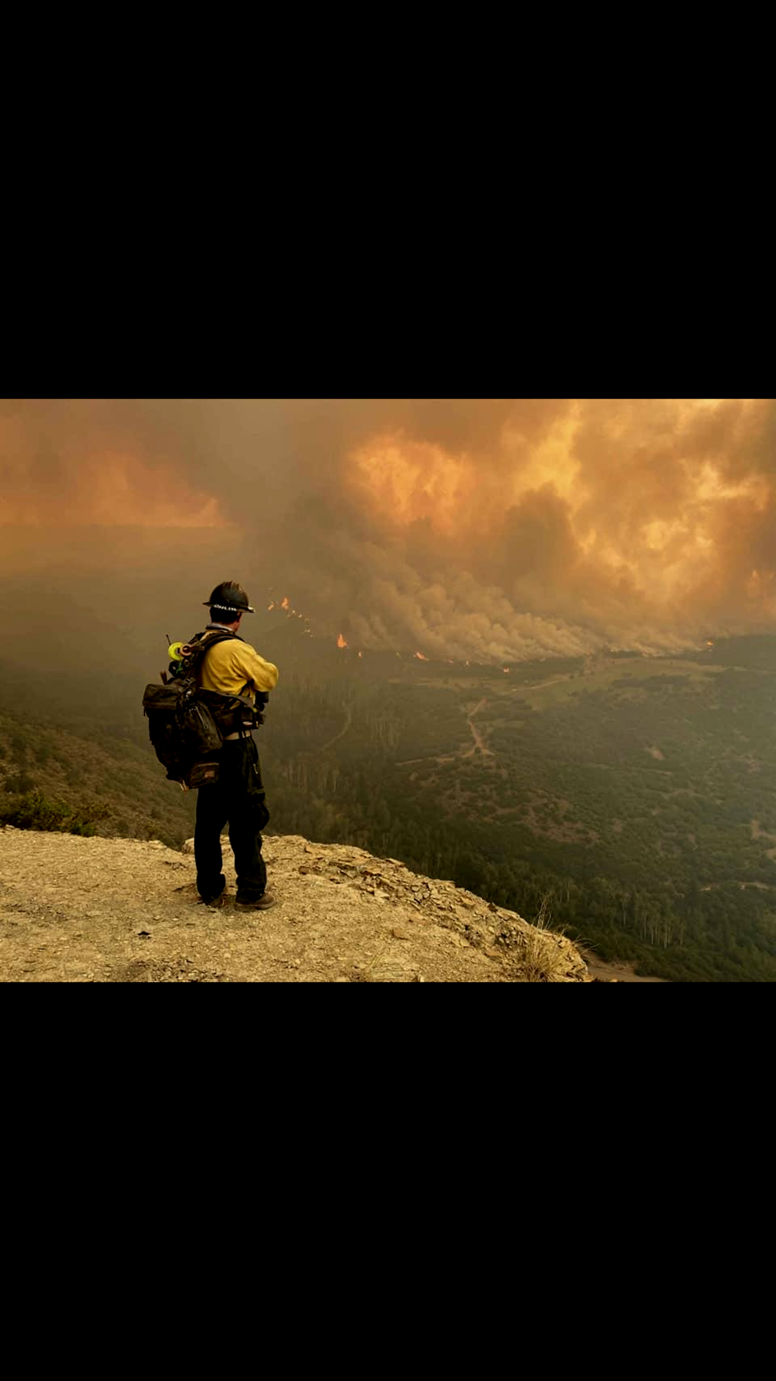 A firefighter overlooks a vast wildfire from a ridge. Smoke fills the sky.