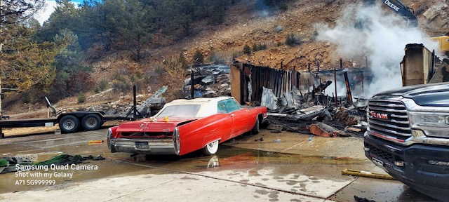 A burnt building, a classic red car, a trailer, and a modern truck, with debris and water on the ground.