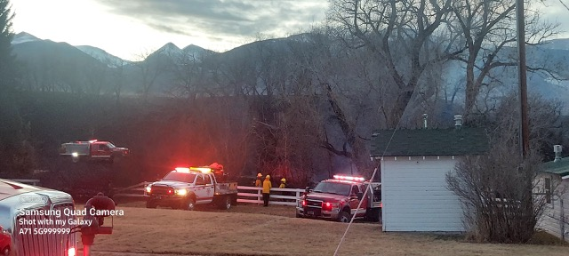 Emergency vehicles with flashing lights and personnel at a scene near a building with mountains in the background at dusk.