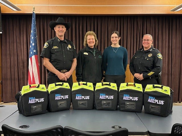 Four individuals in front of an American flag and five defibrillators on a table.