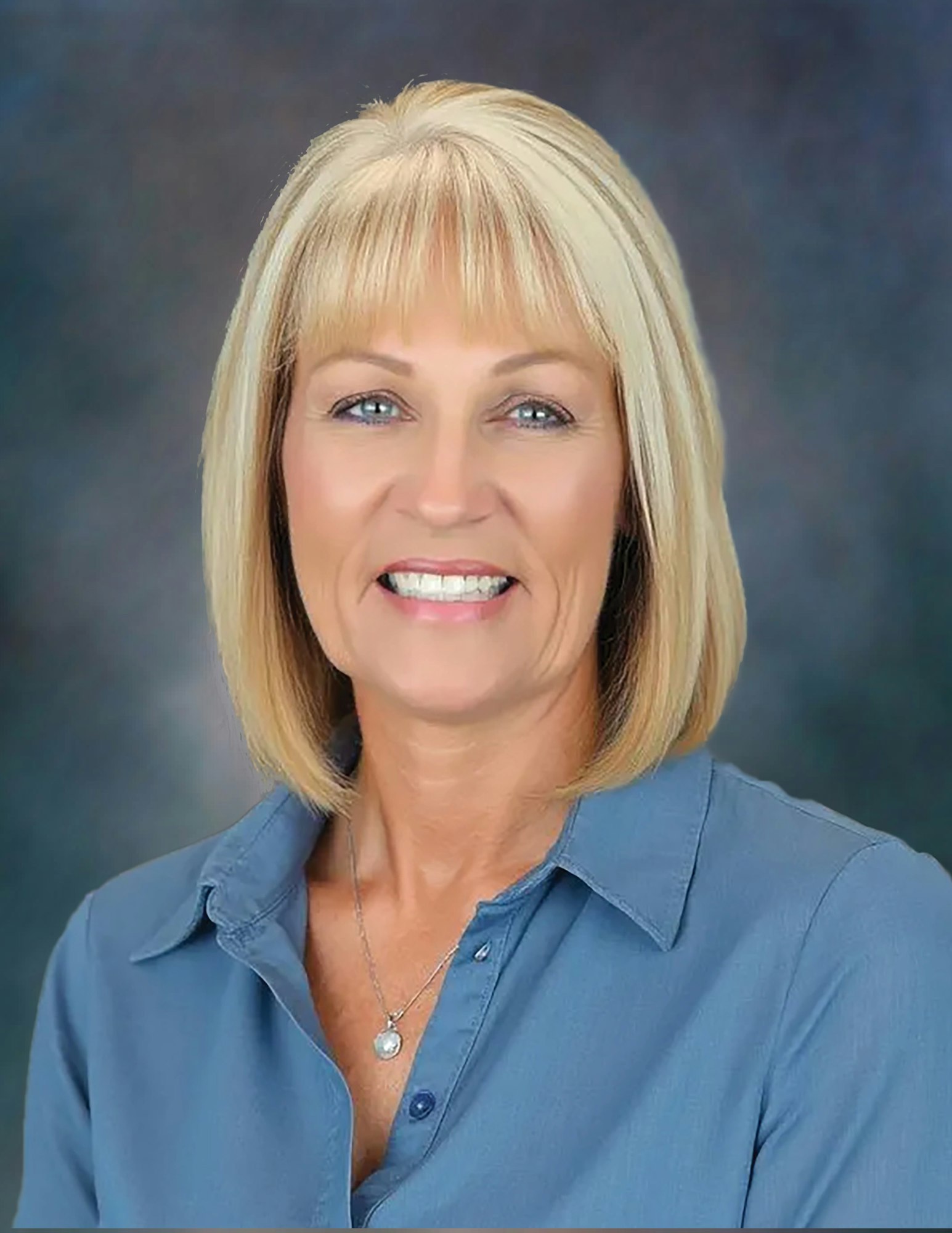 A person with short blonde hair smiles, wearing a blue shirt and a necklace, against a blurred background.