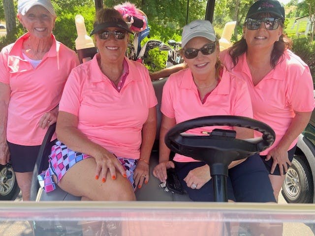 Four women in pink shirts sitting and standing by a golf cart, smiling.