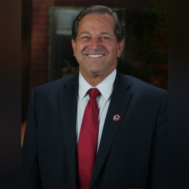 A smiling man in a suit with a red tie and pin, standing before a blurred background.