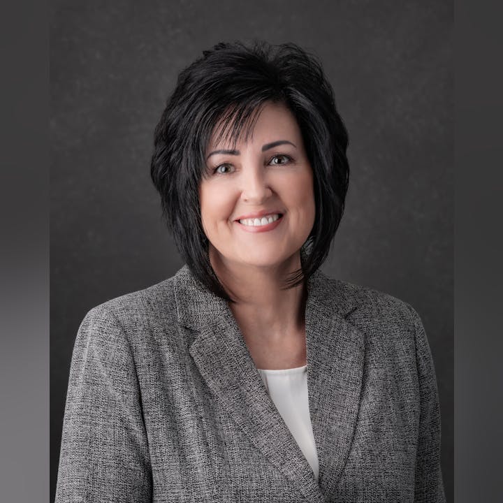 A professional portrait of a smiling woman with black hair in a grey blazer against a grey background.