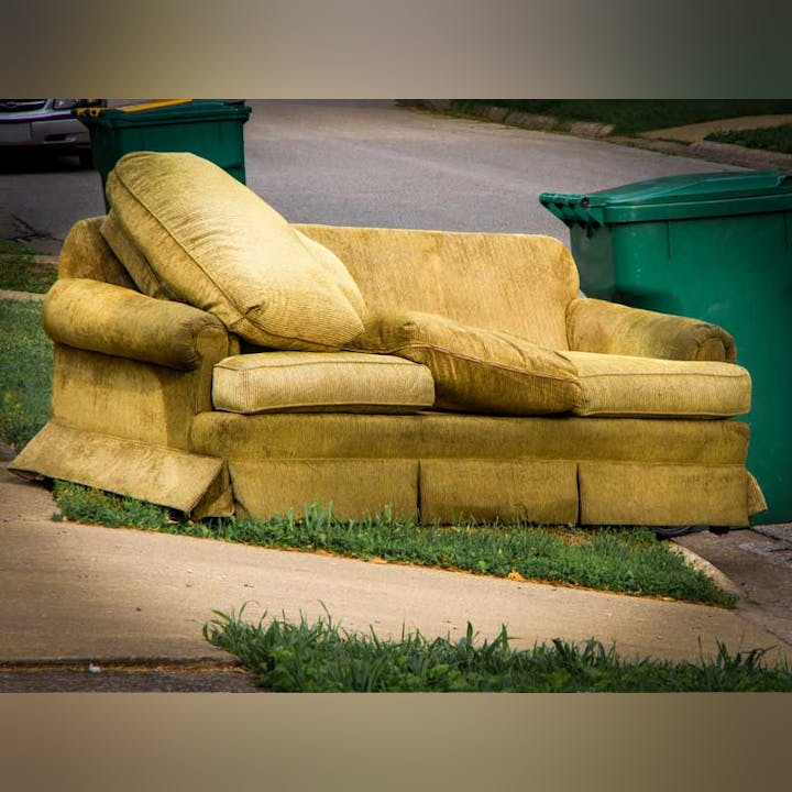 An abandoned yellow couch on the curb next to a green trash bin.