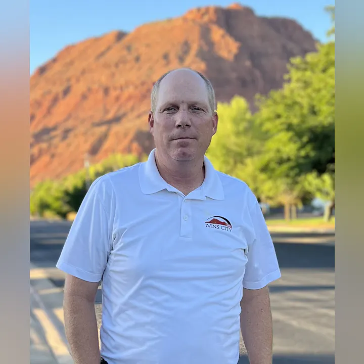 A man in a white polo shirt with "Ivins City" logo, standing in front of a red mountain.