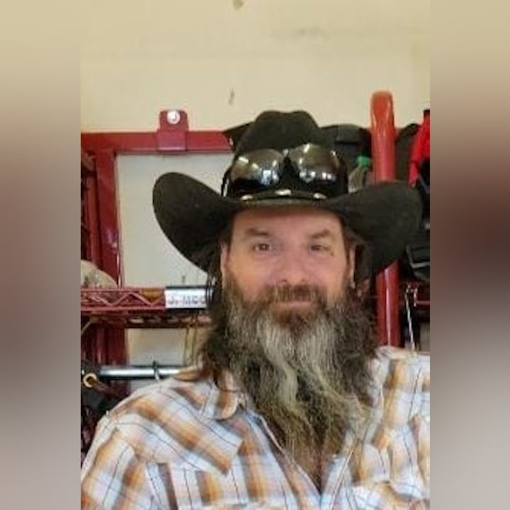 Man with a beard wearing a cowboy hat and sunglasses, smiling in front of a red object.