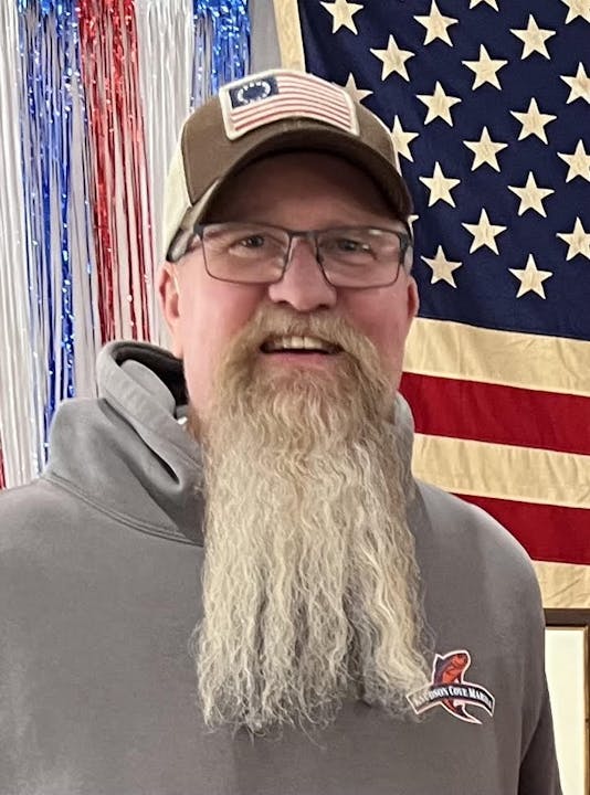 A person with a long gray beard in front of an American flag.