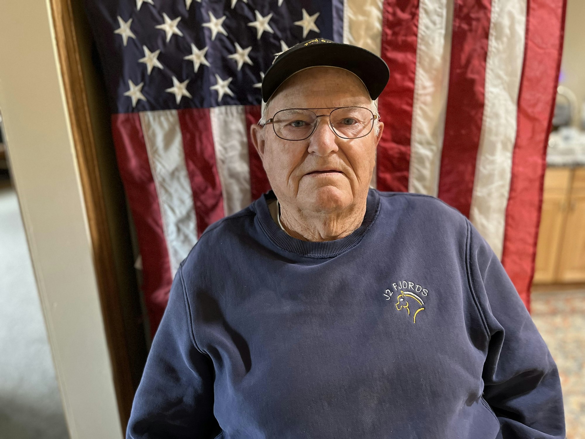 An elderly man wearing glasses and a cap, with a US flag in the background.