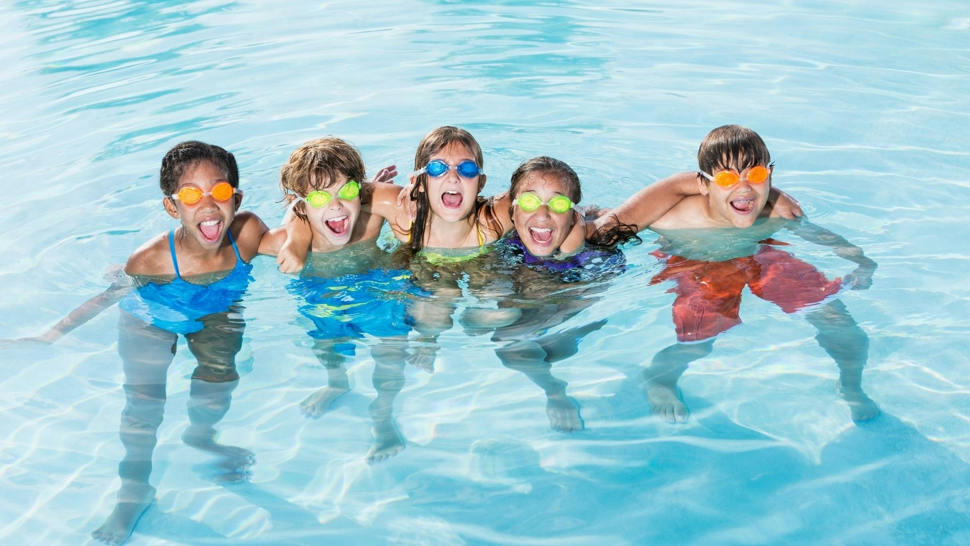 Five kids in goggles, laughing and playing in a swimming pool.