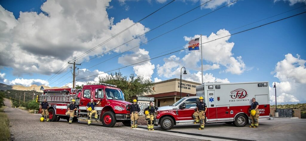 Firefighter crew with firetruck and ambulance outside a station under an American flag.