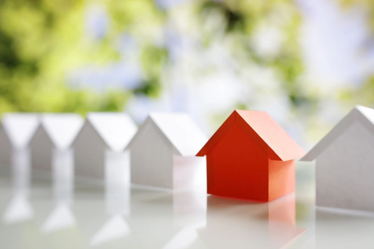 A row of white paper houses with one red house standing out, all on a bright, blurred background.