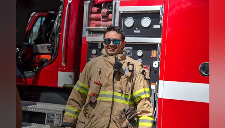 A smiling firefighter in uniform in front of a fire engine.