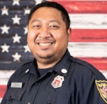 A smiling person wearing a uniform with a badge in front of an American flag.