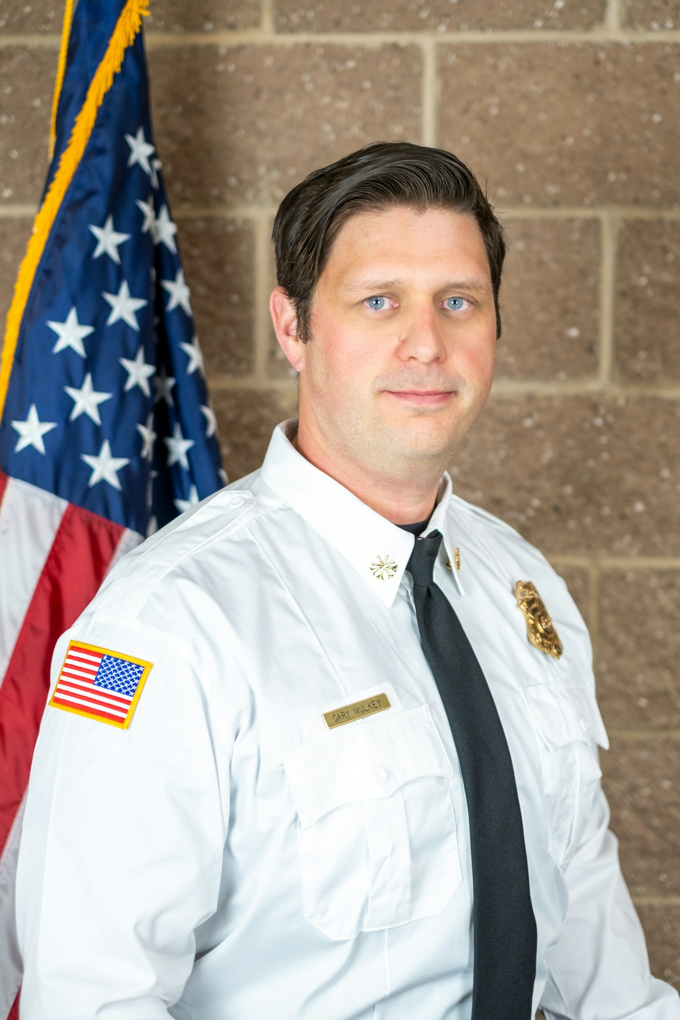 A person in a white uniform with badges and a nametag, standing in front of the U.S. flag, on a brick background.