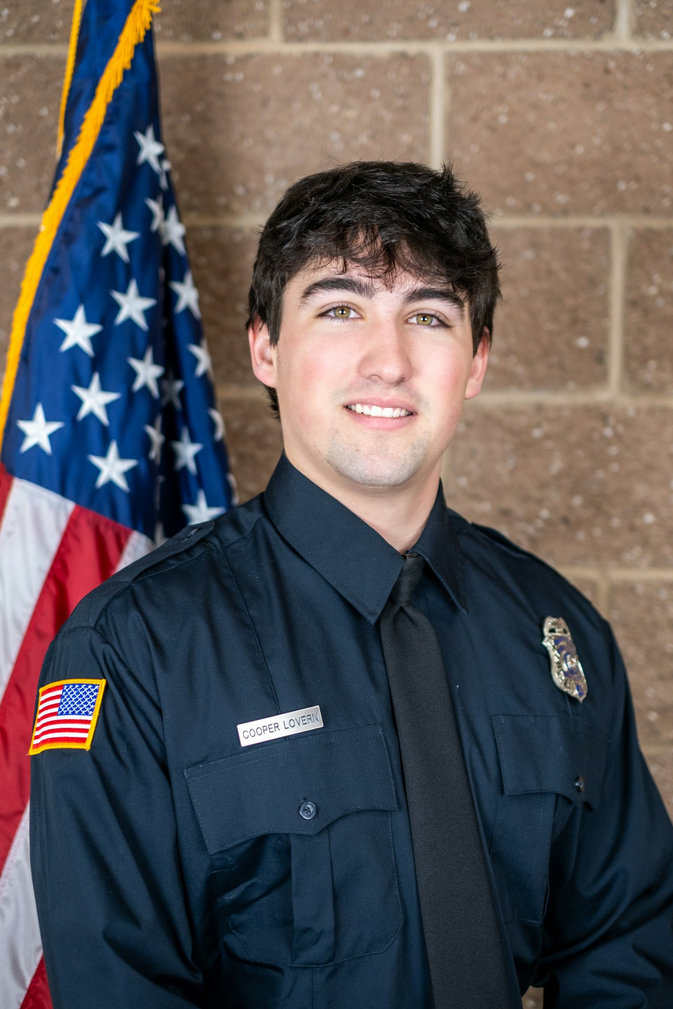 A person in a dark uniform with a name tag, standing in front of a US flag and a brick wall.