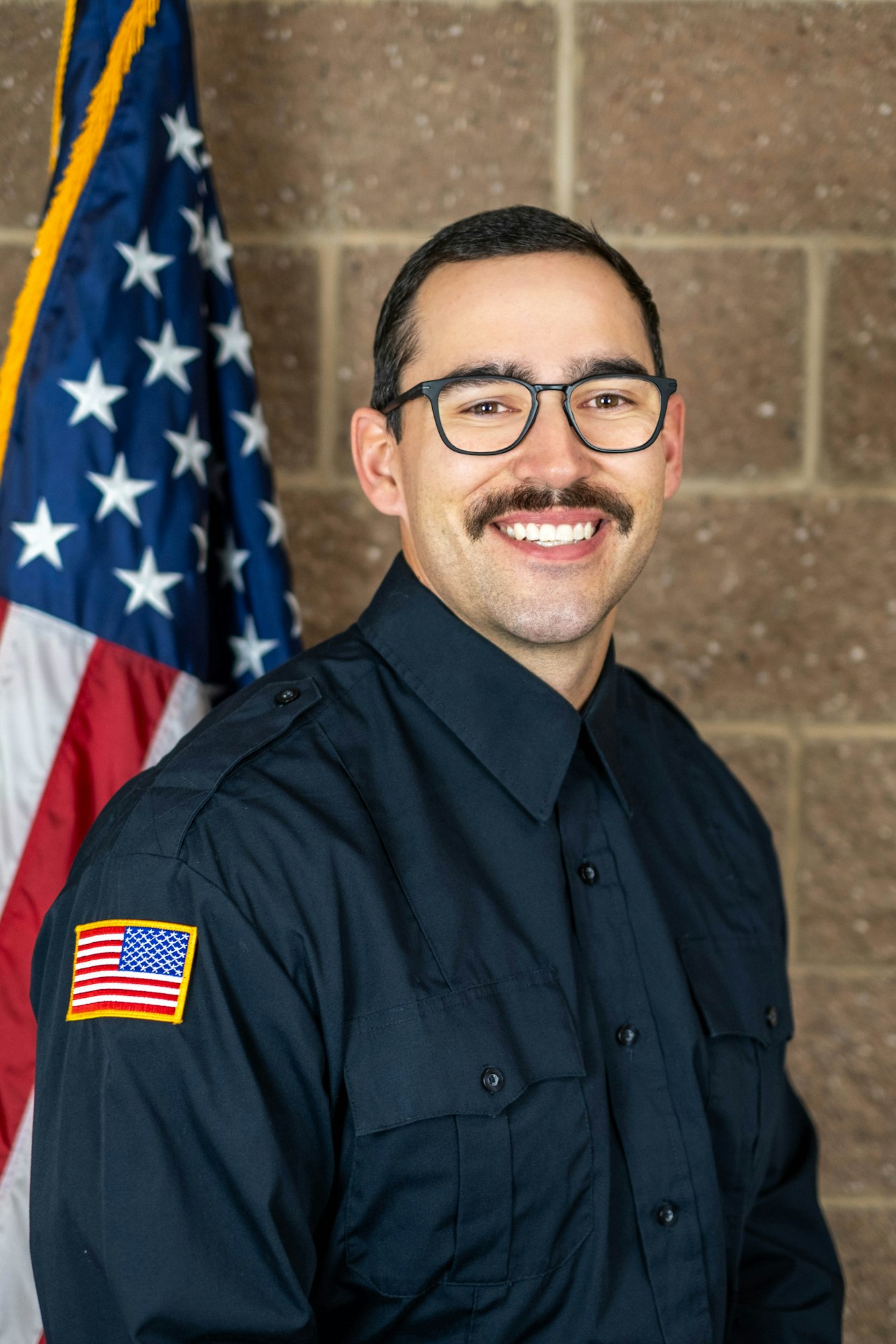 A person in a dark uniform with an American flag patch, standing beside a U.S. flag, smiling, with a brown brick wall background.