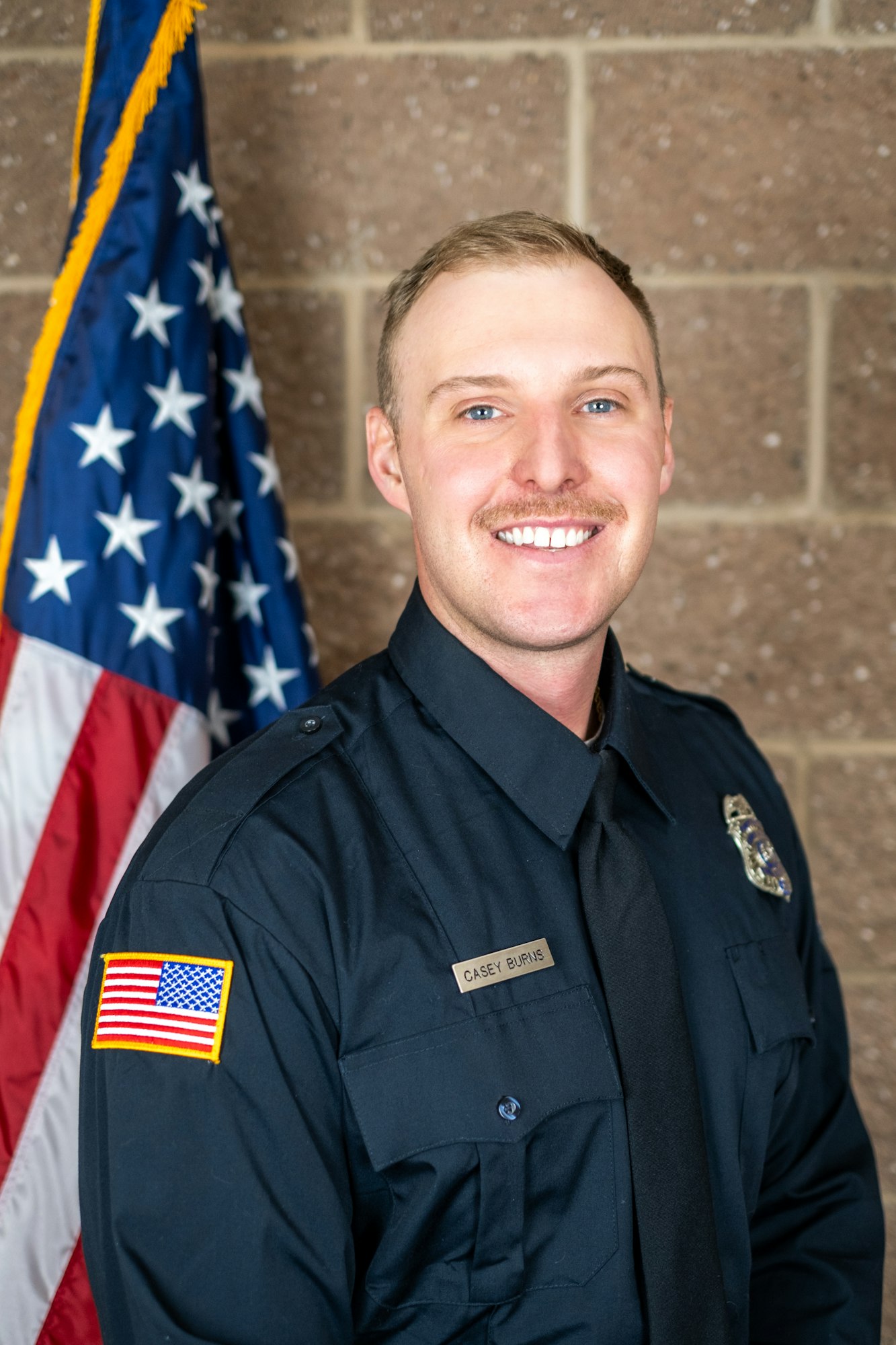 A person in a uniform with badges, standing in front of an American flag, smiling.