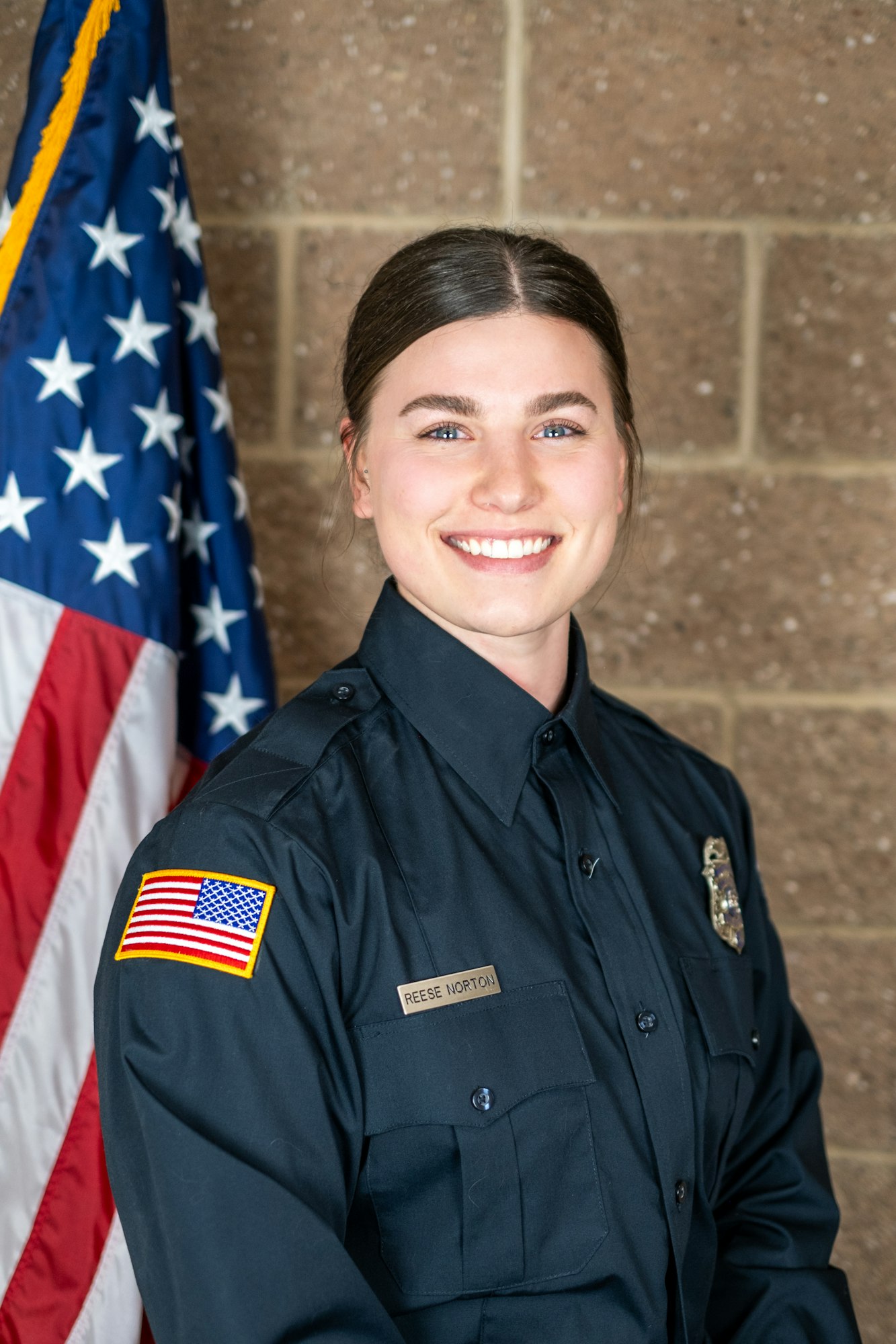 A person in a uniform with a name tag, standing in front of a U.S. flag.