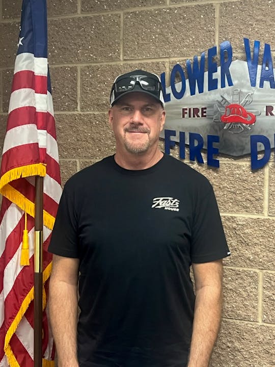 A person in a black shirt and cap stands by an American flag, in front of a wall with "FIRE DEPT." signage.