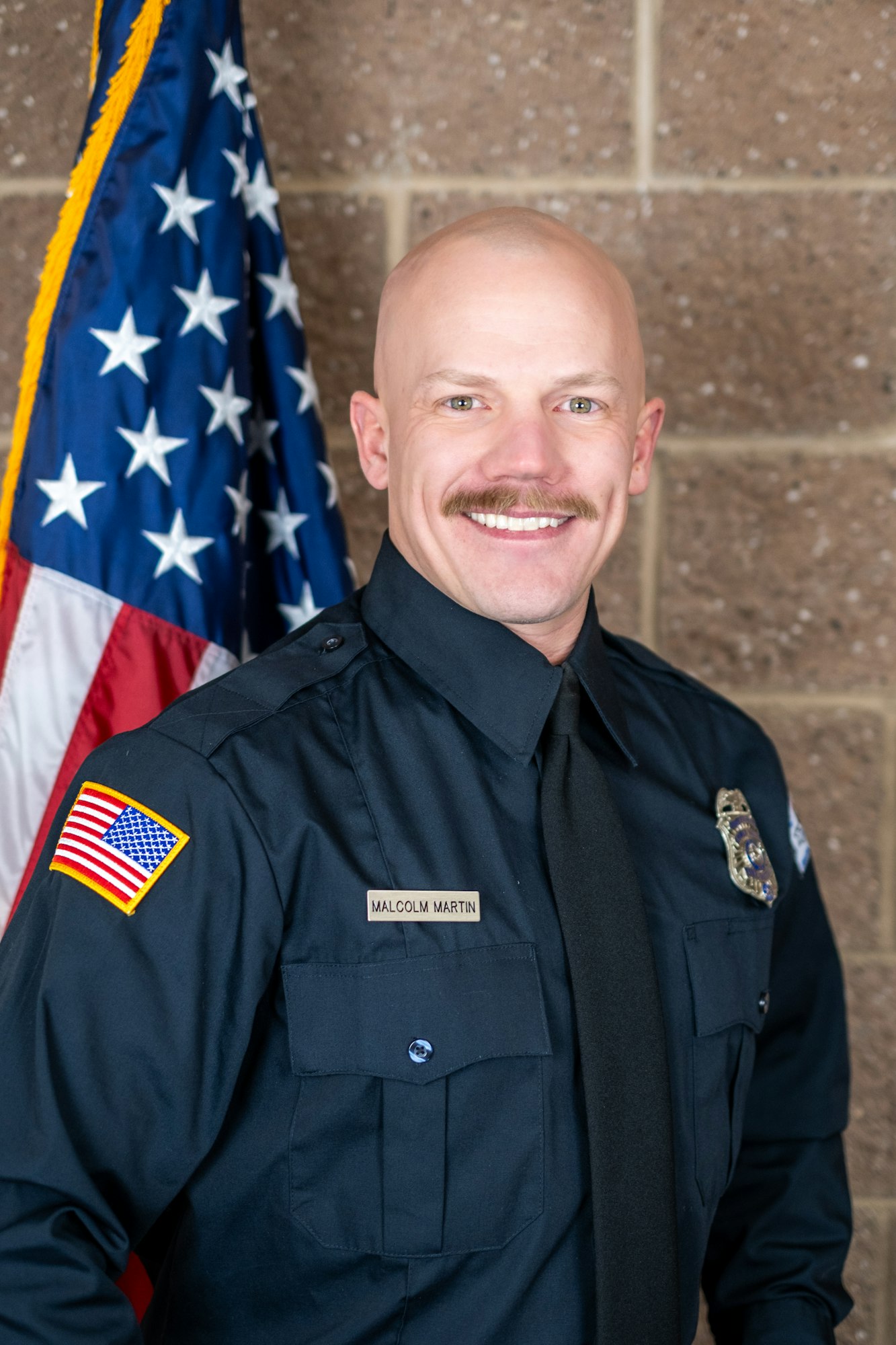 A uniformed officer smiling, standing in front of a U.S. flag, with a badge and name tag visible.