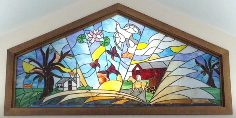 A colorful stained glass window featuring a pastoral scene with birds, a barn, trees, and an open book in the foreground.