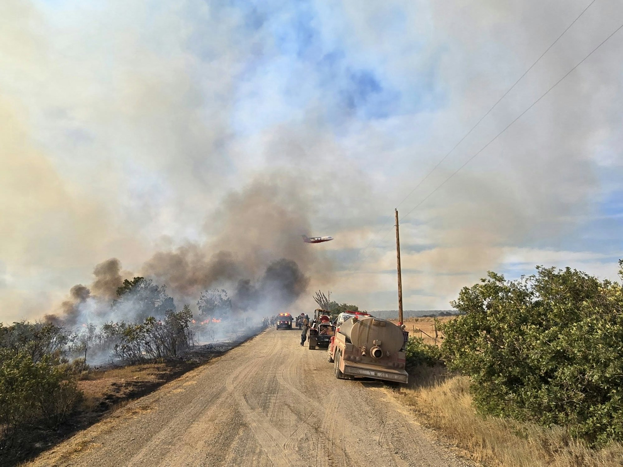 Firefighting efforts on a rural road with smoke in the sky, fire trucks, and a plane above.