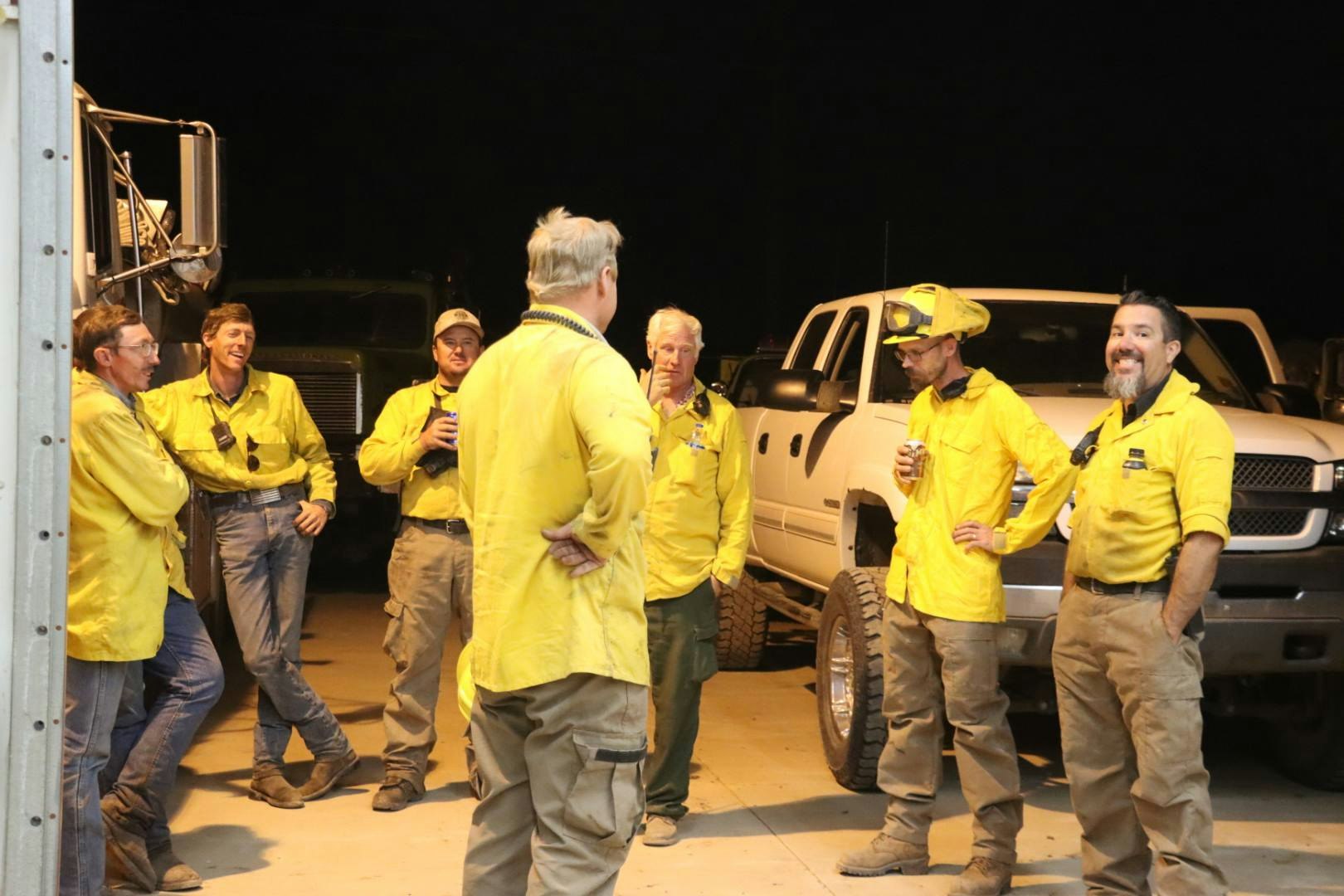 A group of seven people in yellow uniforms, possibly firefighters, are standing and talking near vehicles at night.