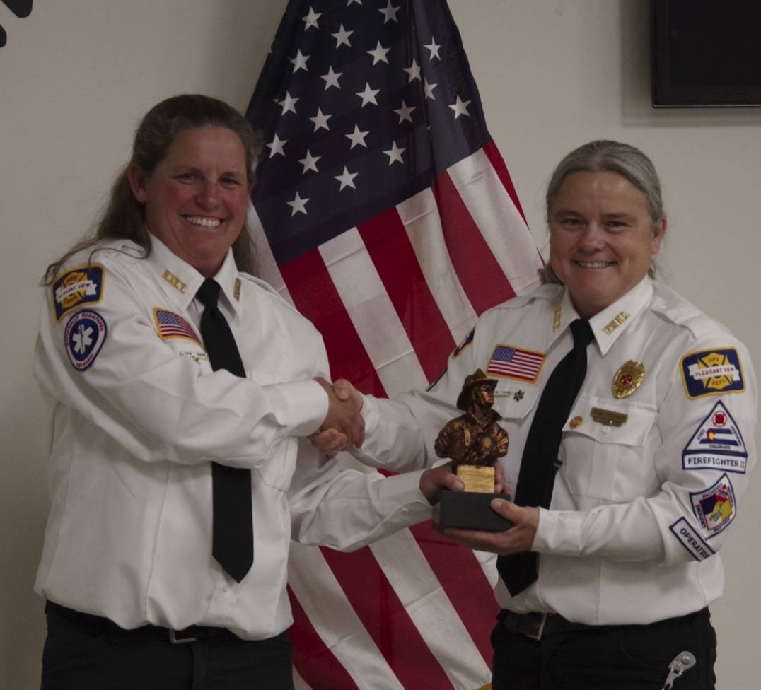 Two emergency service members in uniform shaking hands, one holding an award, with a U.S. flag in the background.