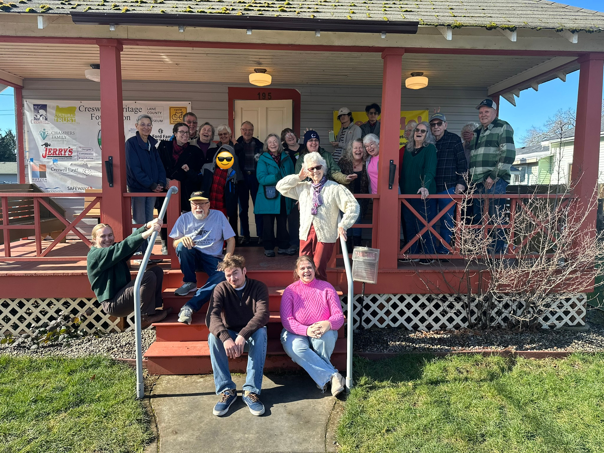 A group of people posing on the porch of a building with various sponsor signs displayed.