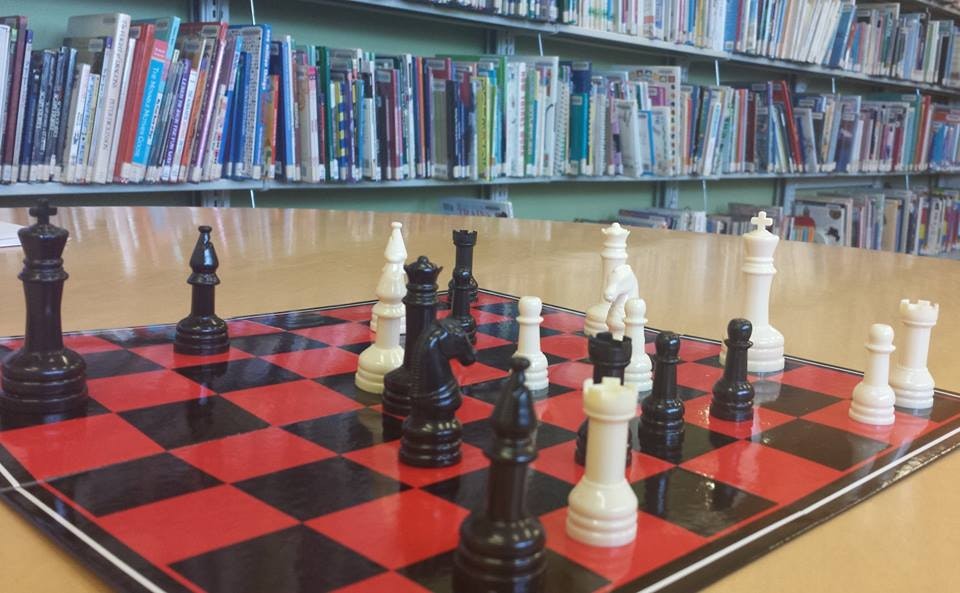 A chessboard with black and white pieces set up, surrounded by bookshelves in a library.