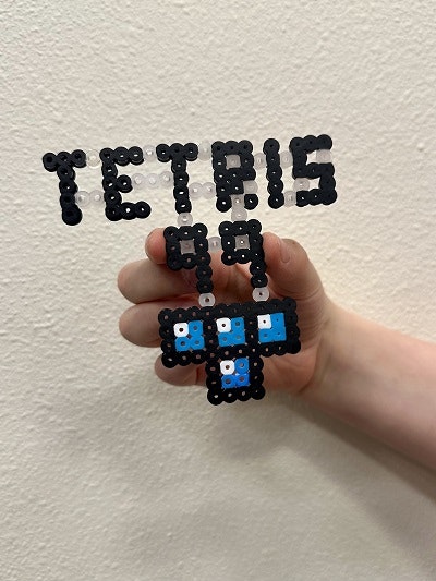 A hand holds a bead art creation that reads "TETRIS 99" with Tetris blocks below.