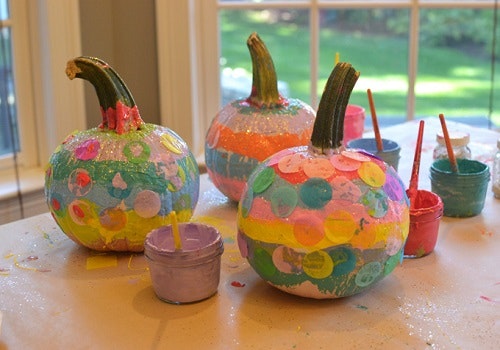 Colorfully painted pumpkins with glitter, paint pots, and brushes on a table, suggesting a craft activity.