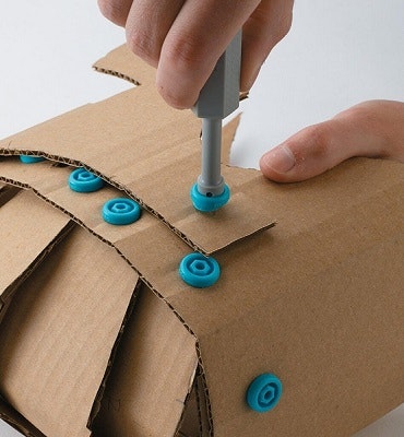 A hand uses a tool to fasten blue plastic rivets on pieces of cardboard.