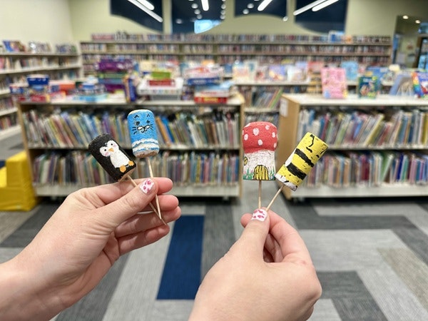 Four painted popsicle sticks resembling animals and characters, held in front of a colorful library backdrop with many books.