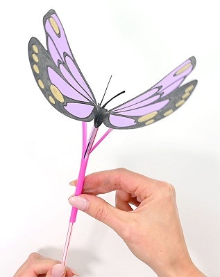 A hand holding a pink stick with a faux purple and yellow butterfly attached, against a white background.