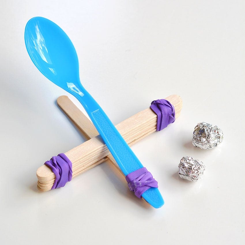 An image featuring a blue plastic spoon resting on two wooden sticks, secured with purple rubber bands, alongside two foil balls.