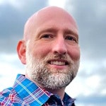 A smiling bald man with a beard in a plaid shirt against a cloudy sky.