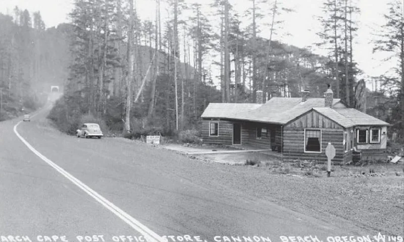 A black and white photo showing a rustic building labeled 'Arch Cape Post Office Store' next to a road with a car passing by.