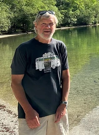 Man standing by a lake, wearing a t-shirt with a camera design, sunglasses on head.