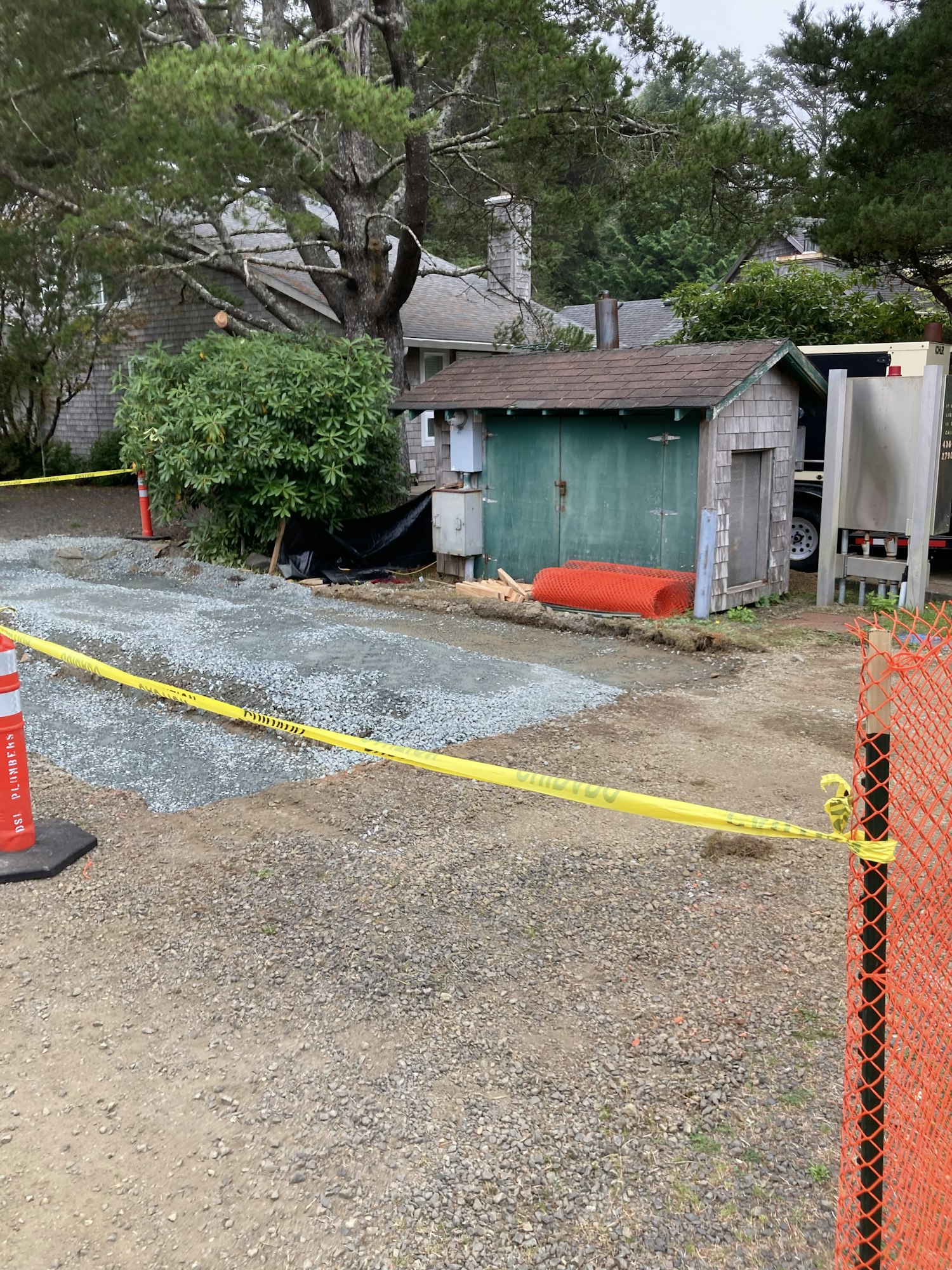 A construction scene with gravel, caution tape, a green shed, and trees.