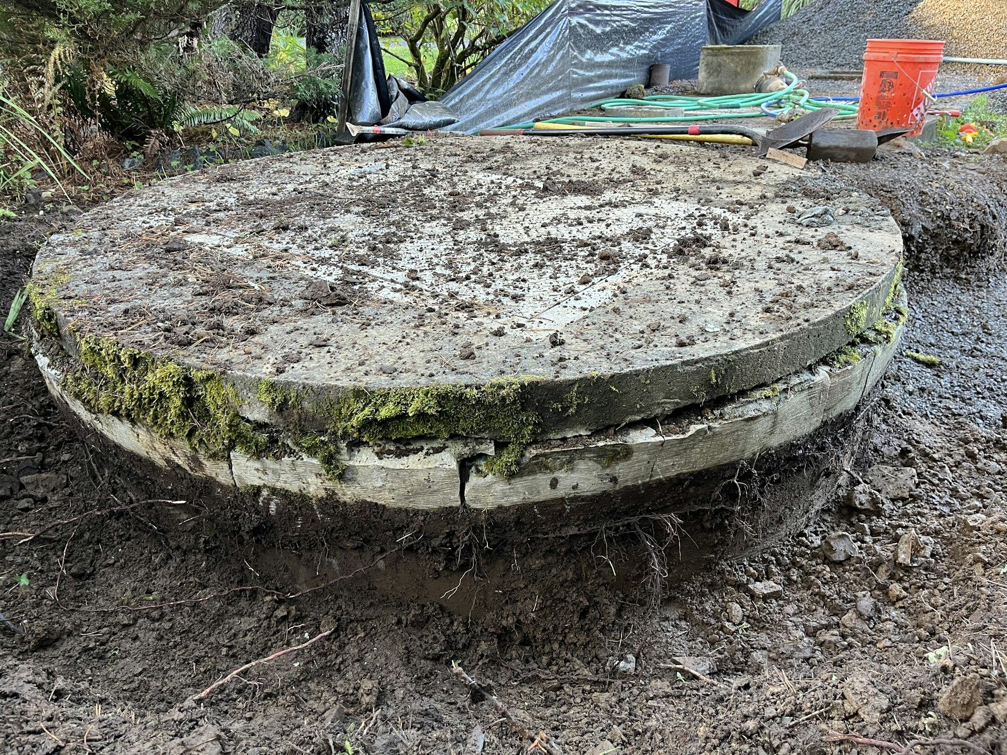 A mossy concrete well cover surrounded by soil and gardening tools.