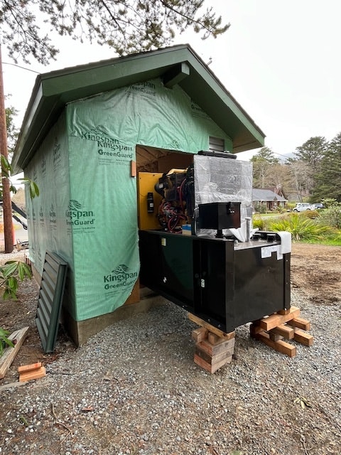 An unfinished shed with a wrapped generator or large machinery on pallets outside.