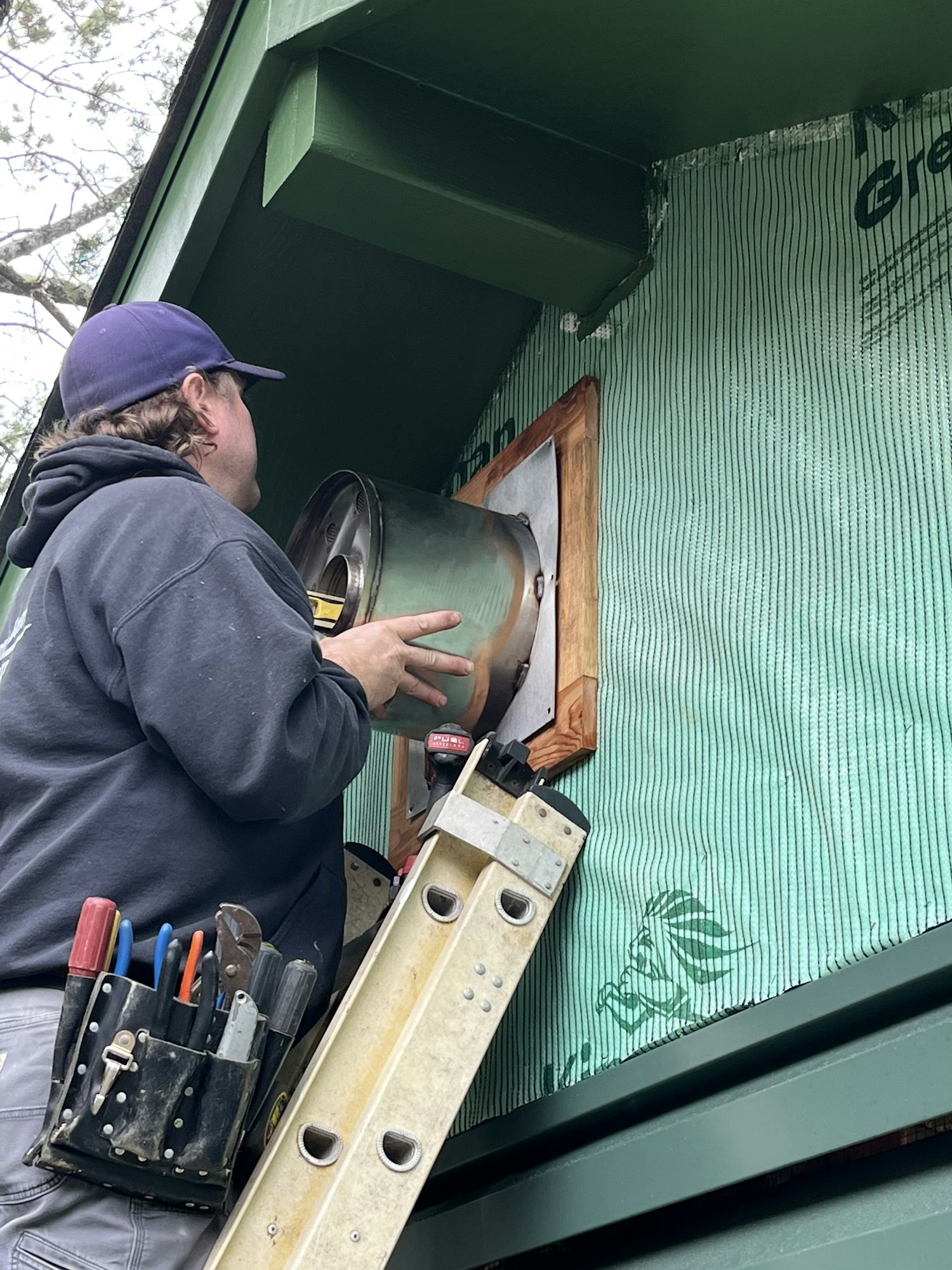 A person is working on a ventilation duct from a ladder, with tools at the ready.