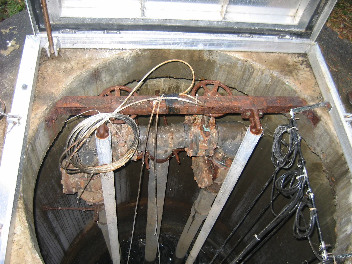 A rusty mechanism, possibly part of a well or underground infrastructure, with cables and pipes visible.