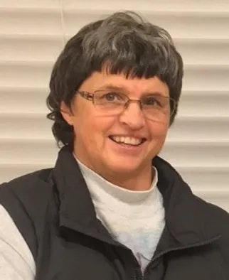 A smiling person with short hair wearing glasses, a vest over a long-sleeved shirt, against a vertical blinds background.